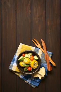 Overhead shot of a bowl of baked vegetables of sweet corn, zucchini, cherry tomato with thyme, toasted bread slice and wooden cutlery on the side, photographed on dark wood with natural light