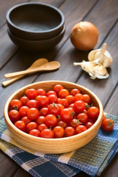 Cherry tomatoes in wooden bowl with garlic, onion, bowls and wooden spoons in the back, photographed on wood with natural light (Selective Focus, Focus one third into the tomatoes)