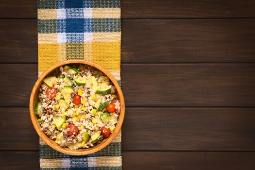 Overhead shot of rice dish with mincemeat and vegetables (sweet corn, cherry tomato, zucchini, onion) in wooden bowl, photographed on dish towel on dark wood with natural light