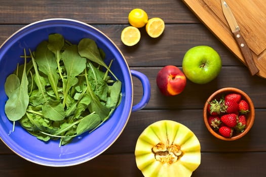 Overhead shot of rucola (lat. Eruca sativa) leaves in blue metal strainer with fresh fruits (lemon, nectarine, apple, strawberry, melon) on the side, photographed on dark wood with natural light