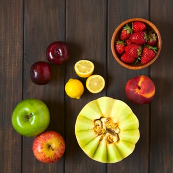 Overhead shot of a variety of fresh fruits (honeydew melon, nectarine, apple, strawberry, plum, lemon), photographed on dark wood with natural light