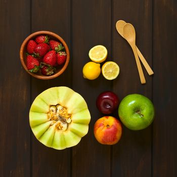 Overhead shot of a variety of fresh fruits (honeydew melon, nectarine, apple, strawberry, plum, lemon) with small wooden spoons on the side, photographed on dark wood with natural light