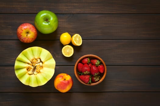 Overhead shot of a variety of fresh fruits (honeydew melon, nectarine, apple, strawberry, plum, lemon), photographed on dark wood with natural light