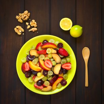 Overhead shot of fresh fruit salad made of grape, strawberry, plum and nectarine served on plate with walnuts, lemon and spoon beside, photographed on dark wood with natural light