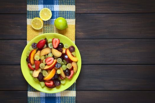 Overhead shot of fresh fruit salad made of grape, strawberry, plum and nectarine served on plate with lemon above, photographed on dark wood with natural light