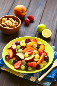 Fresh fruit salad made of grape, strawberry, plum and nectarine served on plate with walnuts, lemon in the back, photographed on dark wood with natural light (Selective Focus, Focus in the middle of the salad)