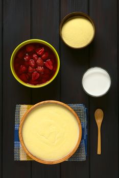 Overhead shot of semolina pudding served in wooden bowl, with a spoon, glass of milk, bowl of strawberry compote, and a bowl of uncooked semolina, photographed on dark wood with natural light  