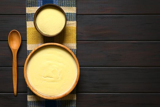 Overhead shot of semolina pudding served in wooden bowl, with a wooden spoon and a bowl of uncooked semolina, photographed on dark wood with natural light  