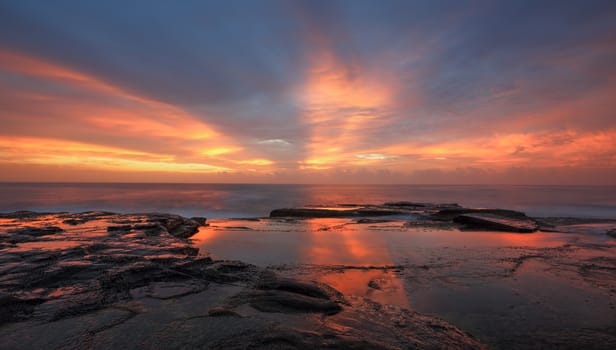 Streaks of orange red light colour the sky and reflect in the water at the rocks of North Curl Curl  Australia