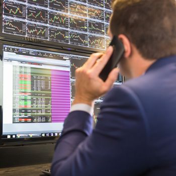 Businessman with cell phone trading stocks. Stock analyst looking at graphs, indexes and numbers on multiple computer screens. Stock trader evaluating economic data.                  