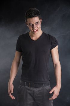 Portrait of a Young Vampire Man with Black T-Shirt, Looking at the Camera, on a Dark Smoky Background.