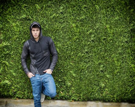 Handsome young man in black hoodie sweater standing outdoor against green plant hedge looking at camera