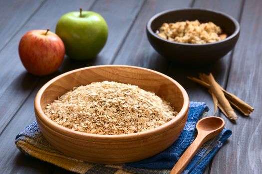 Raw rolled oats in wooden bowl with apples, cinnamon sticks and a bowl of fruit crumble in the back, photographed on dark wood with natural light (Selective Focus, Focus one third into the oats)