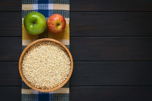 Overhead shot of raw rolled oats in wooden bowl with apples on cloth, photographed on dark wood with natural light