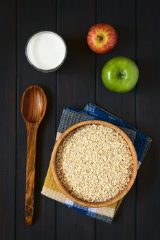 Overhead shot of raw rolled oats in wooden bowl with apples and a glass of milk, photographed on dark wood with natural light