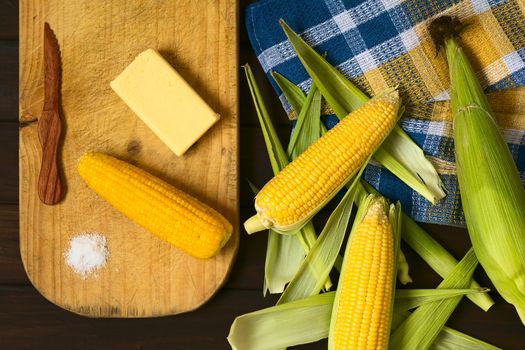 Overhead shot of raw and cooked sweet corn cobs, with butter and salt on wooden board, photographed with natural light