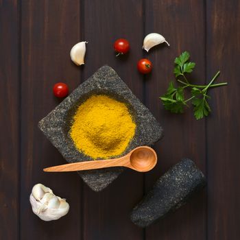 Overhead shot of curry powder spice in mortar with pestle, garlic, cherry tomatoes, parsley leaves on the side, photographed on dark wood with natural light 