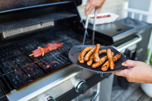 BBQ with sausages and red meat on the grill - male hands holding a plate and taking the meat off the grill before it is too late