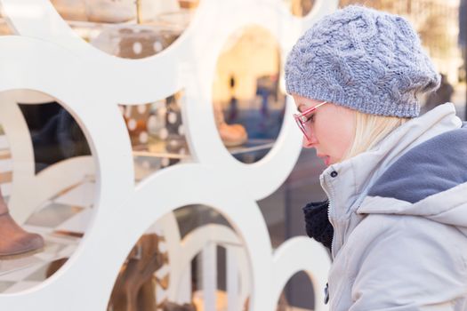 Casualy winter dressed lady window shopping in front of sinfully expensive boutique store dispaly window. Customer woman in shopping street, looking at window, outdoor.