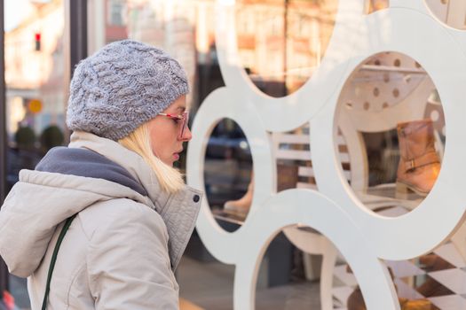 Casualy winter dressed lady window shopping in front of sinfully expensive boutique store dispaly window. Customer woman in shopping street, looking at window, outdoor.