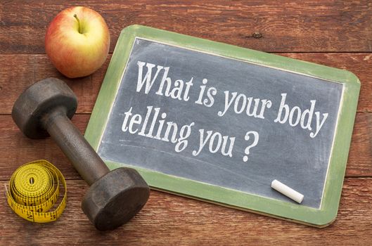What is your body telling you?  A question on a slate blackboard against weathered red painted barn wood with a dumbbell, apple and tape measure
