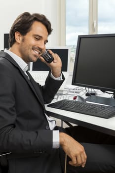 Successful businessman working in his office sitting in a chair at his desk and desktop computer listening to a call on his mobile phone, high angle side view