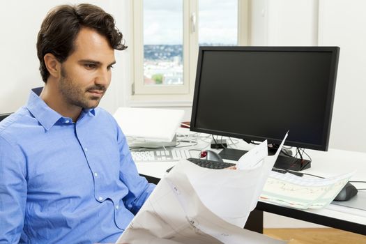 Young handsome man sitting at his desk in the office while reading written agreements and studying important documentation for work