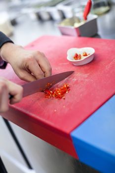 Chef dicing red hot chili peppers on a chopping board in a commercial kitchen for use as a spicy flavouring in his recipes as he cooks the evening meal