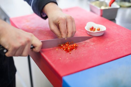 Chef dicing red hot chili peppers on a chopping board in a commercial kitchen for use as a spicy flavouring in his recipes as he cooks the evening meal