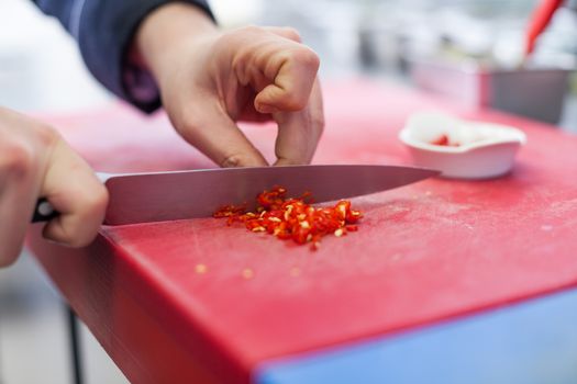 Chef dicing red hot chili peppers on a chopping board in a commercial kitchen for use as a spicy flavouring in his recipes as he cooks the evening meal