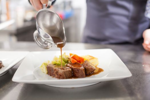 Chef plating up food in a restaurant pouring a gravy or sauce over the meat before serving it to the customer, close up view of his hand and the gravy boat