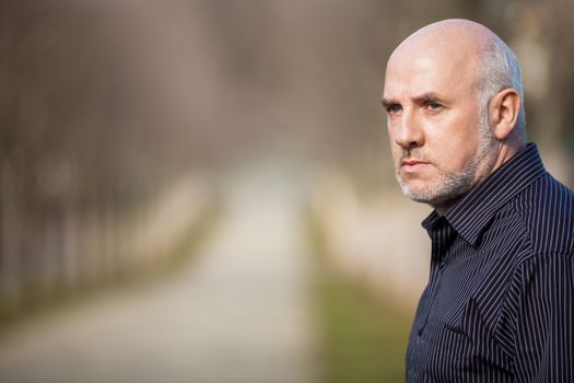 Confident attractive middle-aged man standing waiting in a rural lane with folded arms looking to the right hand side of the frame as though expecting somebody to arrive, with copyspace