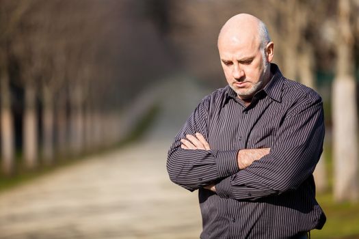 Confident attractive middle-aged man standing waiting in a rural lane with folded arms looking to the right hand side of the frame as though expecting somebody to arrive, with copyspace