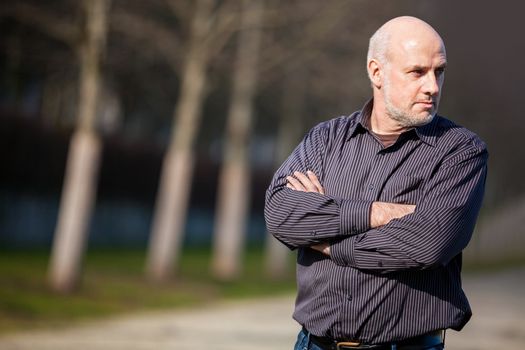 Confident attractive middle-aged man standing waiting in a rural lane with folded arms looking to the right hand side of the frame as though expecting somebody to arrive, with copyspace