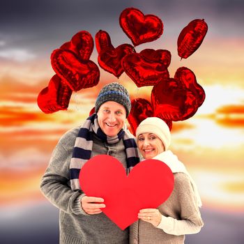 Happy mature couple in winter clothes holding red heart against purple sky with orange clouds
