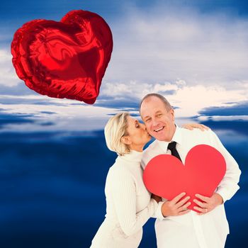 Older affectionate couple holding red heart shape against blue sky with blue clouds