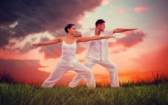 Peaceful couple in white doing yoga together in warrior position against red sky over grass