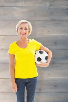 Mature blonde holding football smiling at camera against pale grey wooden planks