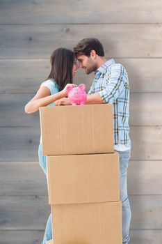 Happy young couple with moving boxes and piggy bank against pale grey wooden planks