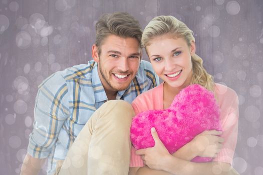 Attractive young couple sitting holding heart cushion against grey abstract light spot design