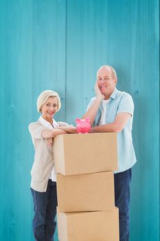 Older couple smiling at camera with moving boxes and piggy bank against wooden planks background