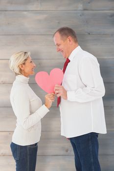 Older affectionate couple holding pink heart shape against pale grey wooden planks