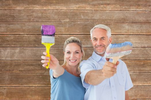 Happy couple holding paintbrushes smiling at camera against wooden planks background
