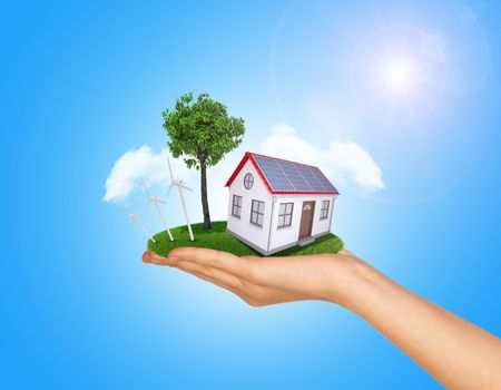 Hand holding house on green grass with tree, wind turbine and solar panels. Background clouds and blue sky