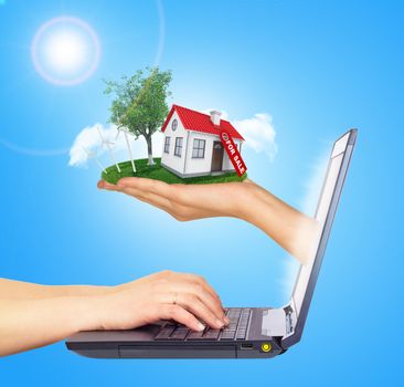 White shack in hand with red roof, brown door and chimney of screen laptop. Hands typing on keyboard. Background sun shines brightly on left. Blue sky