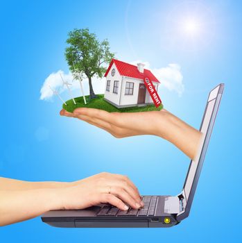 White shack in hand with red roof, brown door and chimney of screen laptop. Hands typing on keyboard. Background sun shines brightly on right. Blue sky