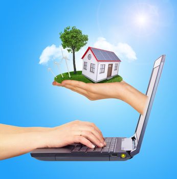 Hand holding house on green grass with tree, wind turbine and solar panels of screen laptop. Hands typing on keyboard. Background clouds and blue sky