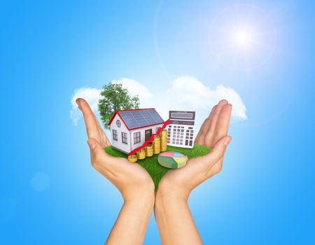 Hands holding green grass with house on ground. Standing tree and clouds in background. Blue isolated