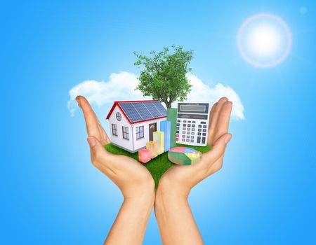 Hands holding green grass with house on ground. Standing tree and clouds in background. Blue isolated