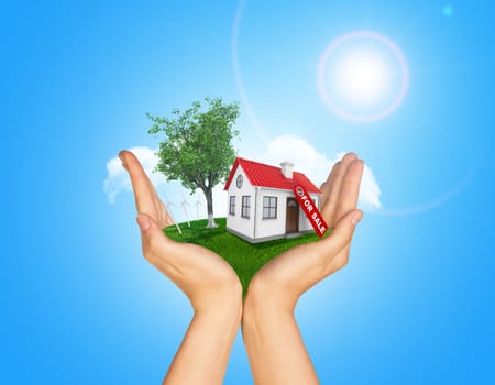 Hands holding house on green grass with label for sale, red roof, chimney, tree, wind turbine. Background clouds and blue sky